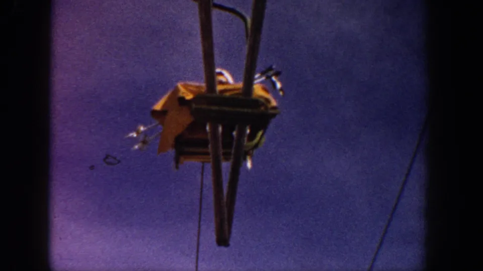 Close-up detail of a vintage single-seat ski lift grip assembly and cable hardware, yellow chair visible from below against a blue-purple sky, circa 1961.