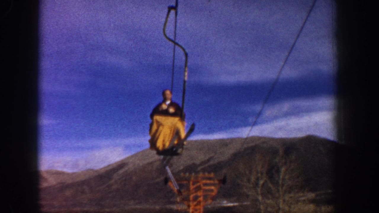 Man on chairlift in yellow-orange jacket at higher elevation; orange lift tower, bare trees, and mountain ridgeline visible behind.