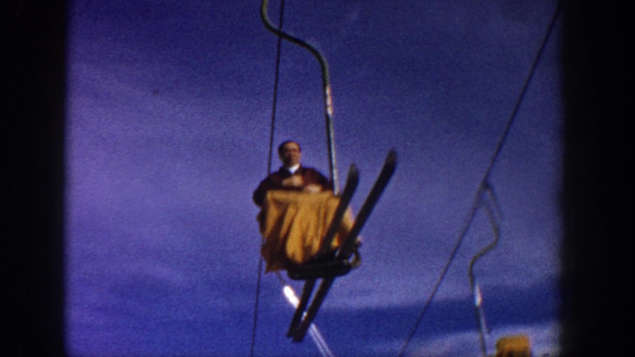Man in yellow-orange jacket on open chairlift with skis angled downward; blue sky and mountain backdrop, circa 1961 Aspen, Colorado, 8mm.