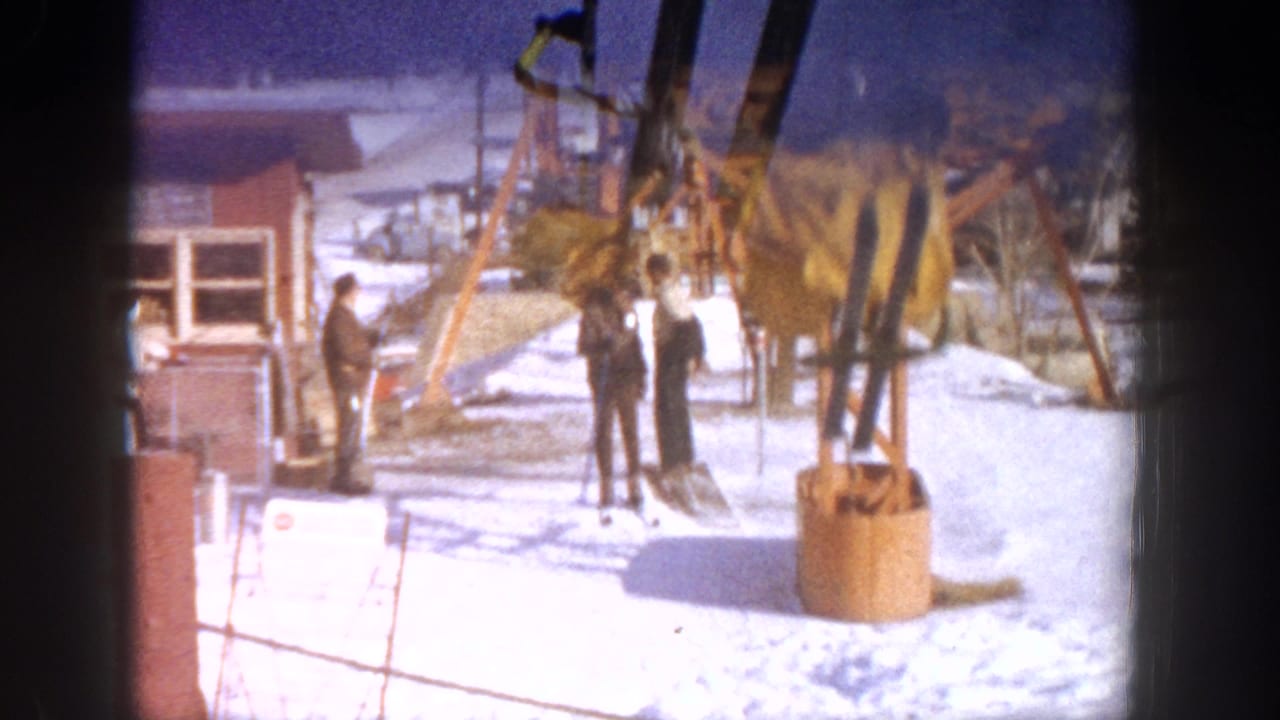 Ski lift base station at Aspen, Colorado, circa 1961; several figures in winter dress near orange metal lift mechanism and red-brown structure on 8mm film.