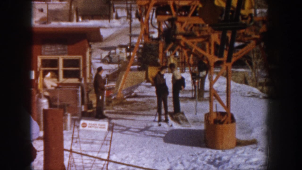 Several adults in winter coats stand near an orange steel chairlift tower base at what is described as Aspen, Colorado, in 1961, on a bright winter day.