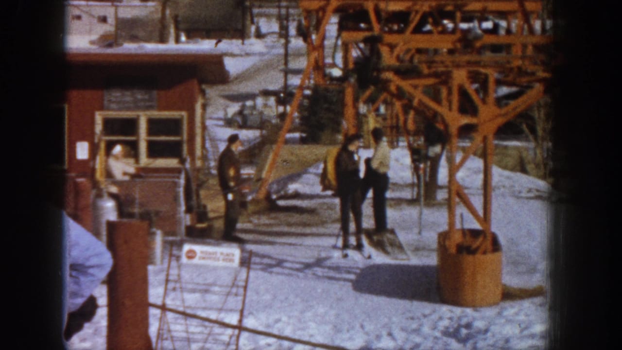 Adults in winter clothing stand near orange lift tower machinery and a wooden structure at a snowy ski resort base.