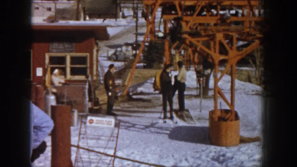 A group of adults in mid-century winter attire congregates near an orange ski lift tower and wooden lodge building at what appears to be Aspen, Colorado, circa 1961.