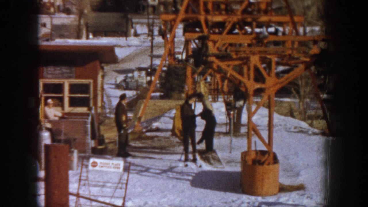 A skier appears to be removing a coat near the orange lift tower base at a Colorado ski resort, circa 1961, with other visitors and the wooden building visible behind.
