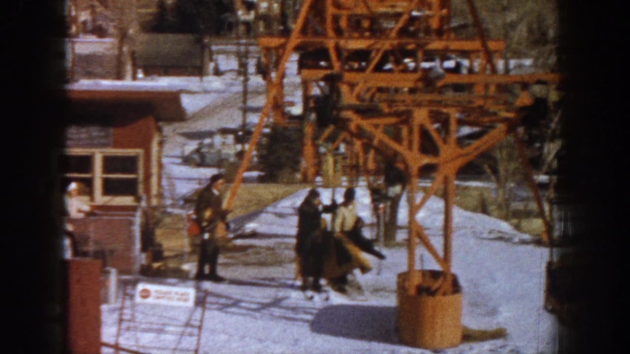 End frame showing skiers gathered around the orange chairlift tower base at an early-1960s Colorado ski resort, wooden building visible at left, winter daylight overhead.