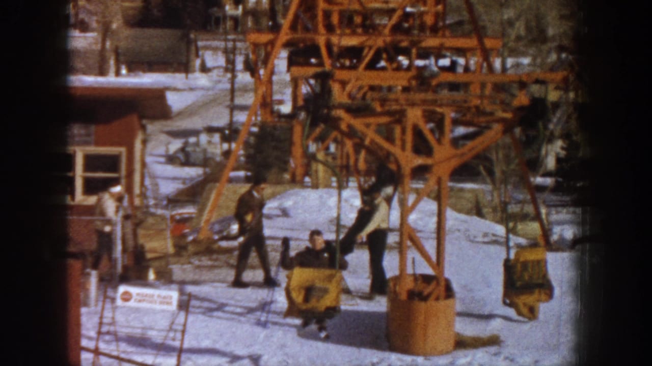Skiers in dark winter coats near an orange wooden ski lift tower on snow, with a red resort building behind, 8mm film.