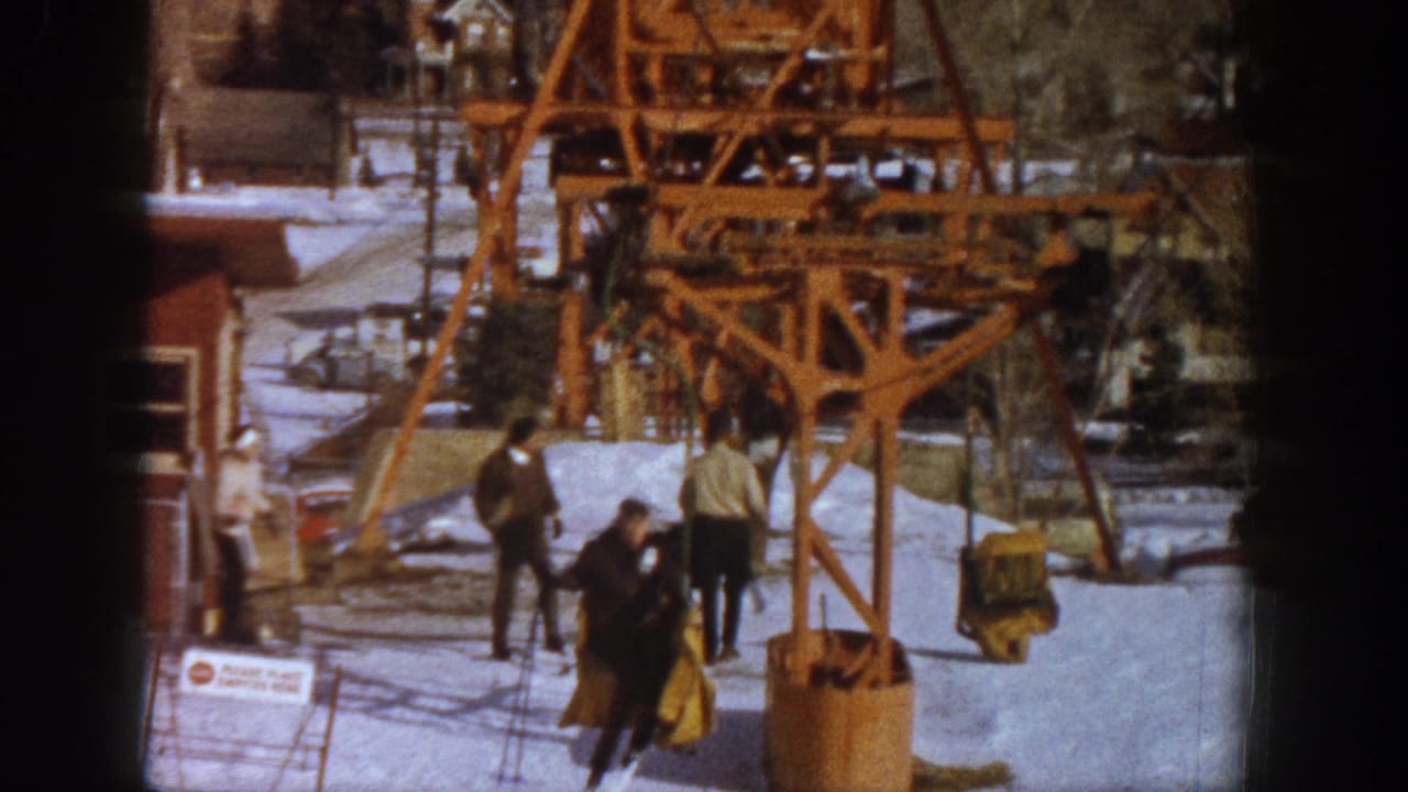 8mm home movie frame of skiers gathering near a large wooden lift structure, with a red resort building visible in background, Aspen ski resort, Colorado, circa 1961.