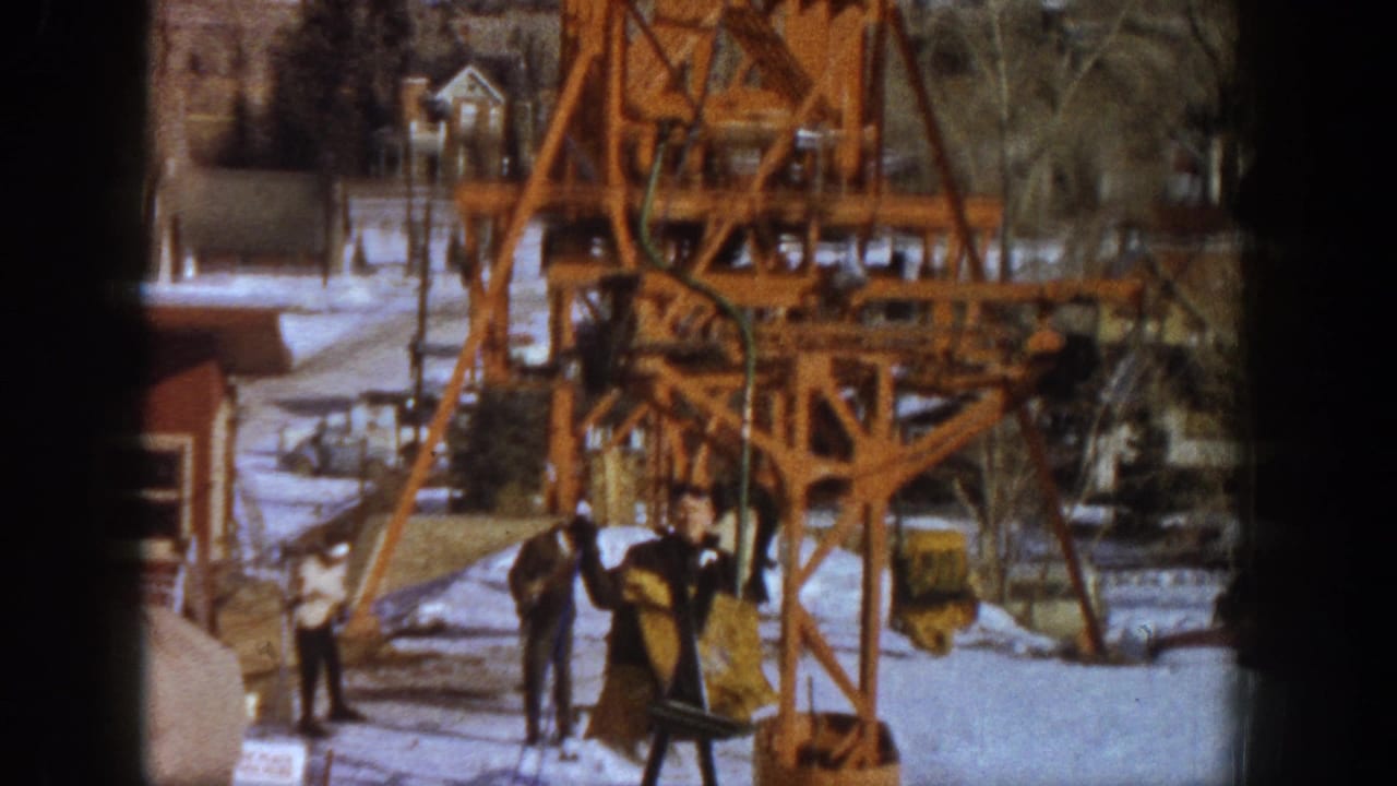 1961 Aspen, Colorado — a skier in dark jacket and yellow pants rides an orange lattice-steel lift while visitors stand in snow below.