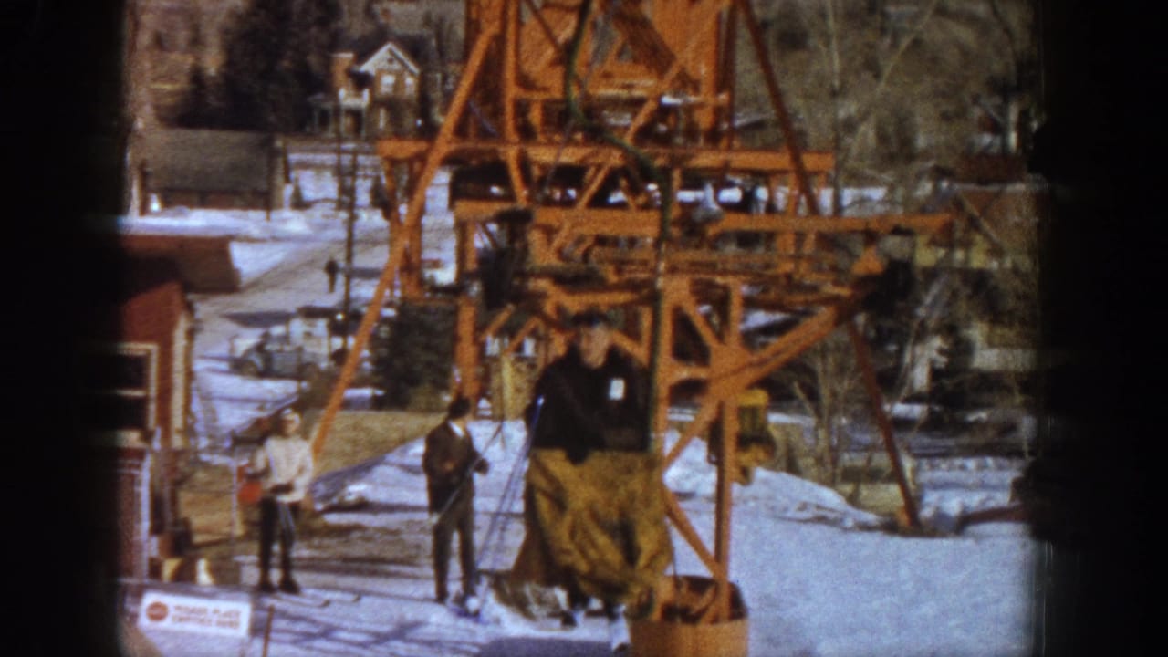 Orange ski lift structure; rider in dark jacket and yellow pants on the lift; other visitors and snow-covered buildings in background.