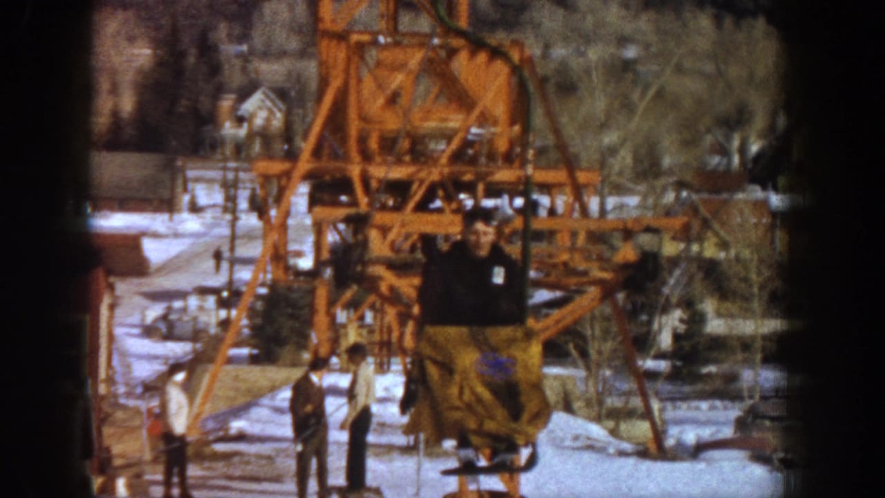 1961 Aspen, Colorado — a lift rider in dark jacket with yellow pants against the orange steel structure; a second person stands at lower left in the snow.