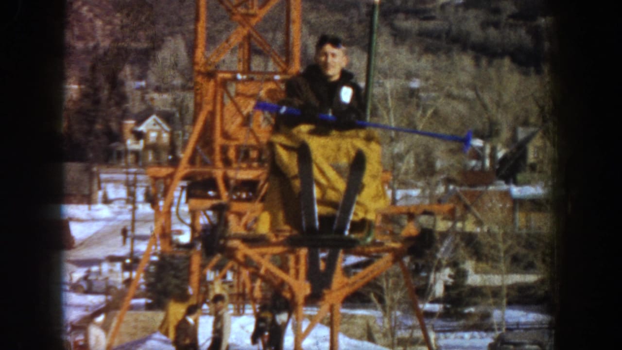 1961 Aspen, Colorado — final frame showing the orange lift structure with a rider in dark jacket and yellow pants; snow-covered resort buildings behind.
