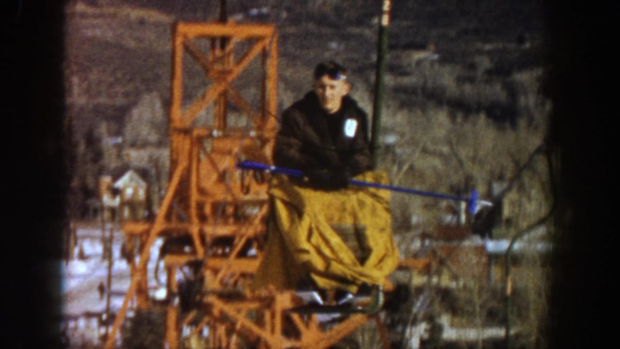 Skier riding orange lattice chairlift at Aspen, Colorado, c.1961; dark jacket, yellow trousers, blue ski poles; bare winter trees and valley buildings below.
