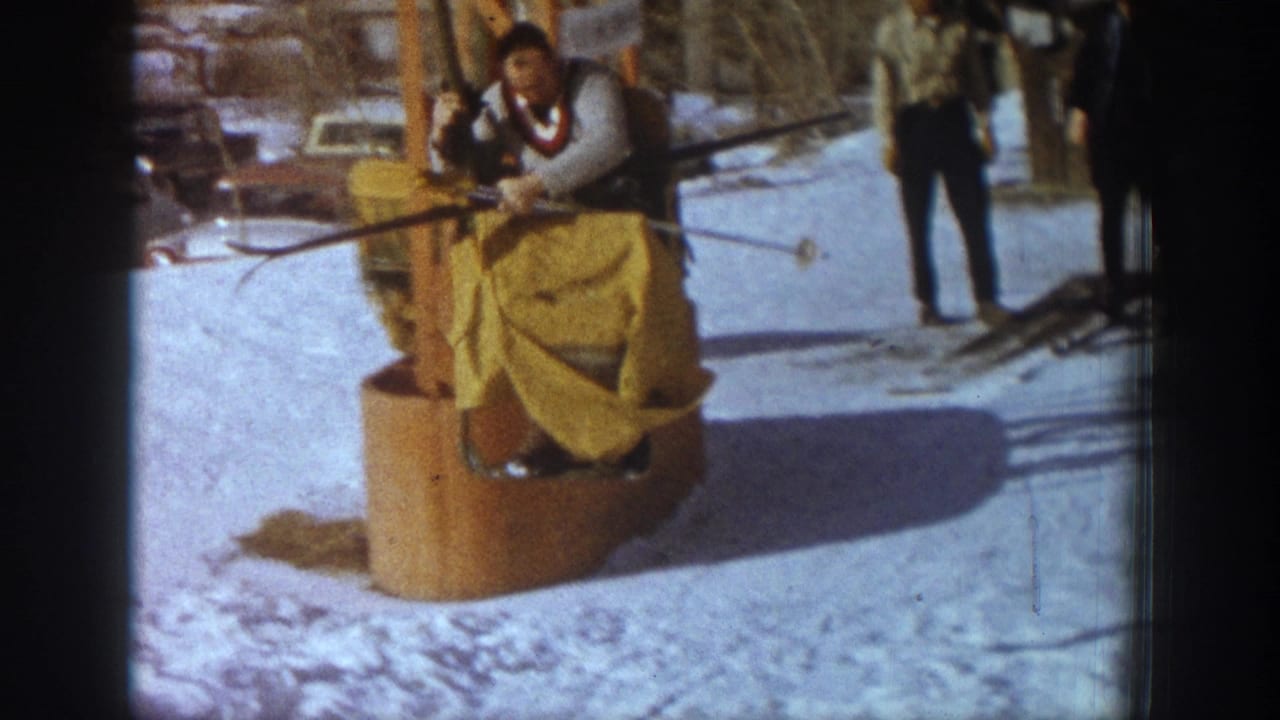 Close view of skier arriving at chairlift bottom terminal; bystander in dark clothing at right; orange chairlift seat and tower prominent; background vehicles on snow.
