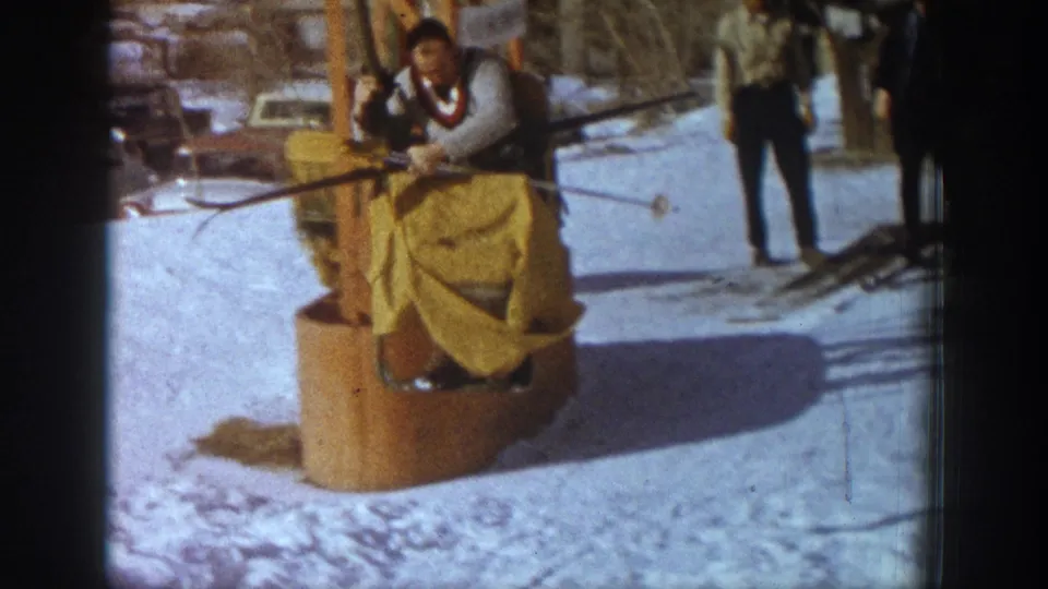 Skier arriving at Aspen chairlift bottom terminal, c.1961; yellow fabric draped over lap, colorful neckwear visible; standing bystander at right; vehicles in background.