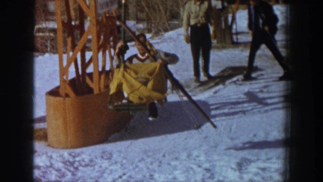 Skier disembarking from orange chairlift terminal at Aspen ski resort, c.1961; yellow fabric garment visible as rider slides onto snow; two figures in dark attire nearby.