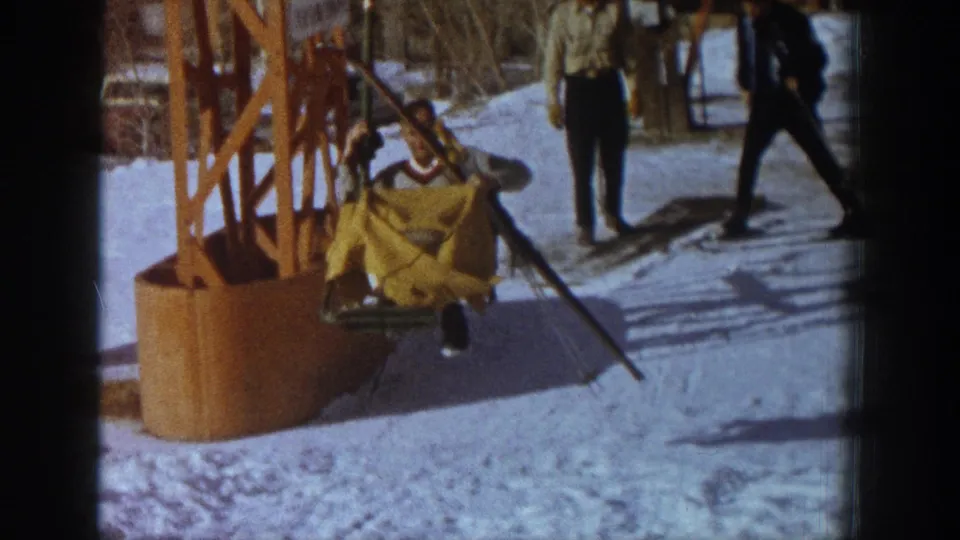 Skier disembarking from orange chairlift terminal at Aspen ski resort, c.1961; yellow fabric garment visible as rider slides onto snow; two figures in dark attire nearby.