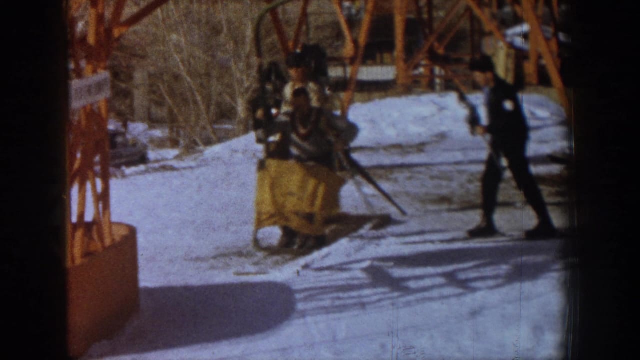 Skier on snow after disembarking at Aspen chairlift terminal, c.1961; second figure in dark ski attire at right; orange lift tower and bare trees in background.
