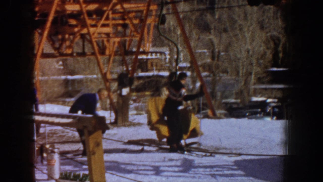 Close view of rust-orange ski lift tower framework at a 1960s mountain resort, likely Aspen, with yellow-jacketed skier and others preparing to board in warm 8mm tones