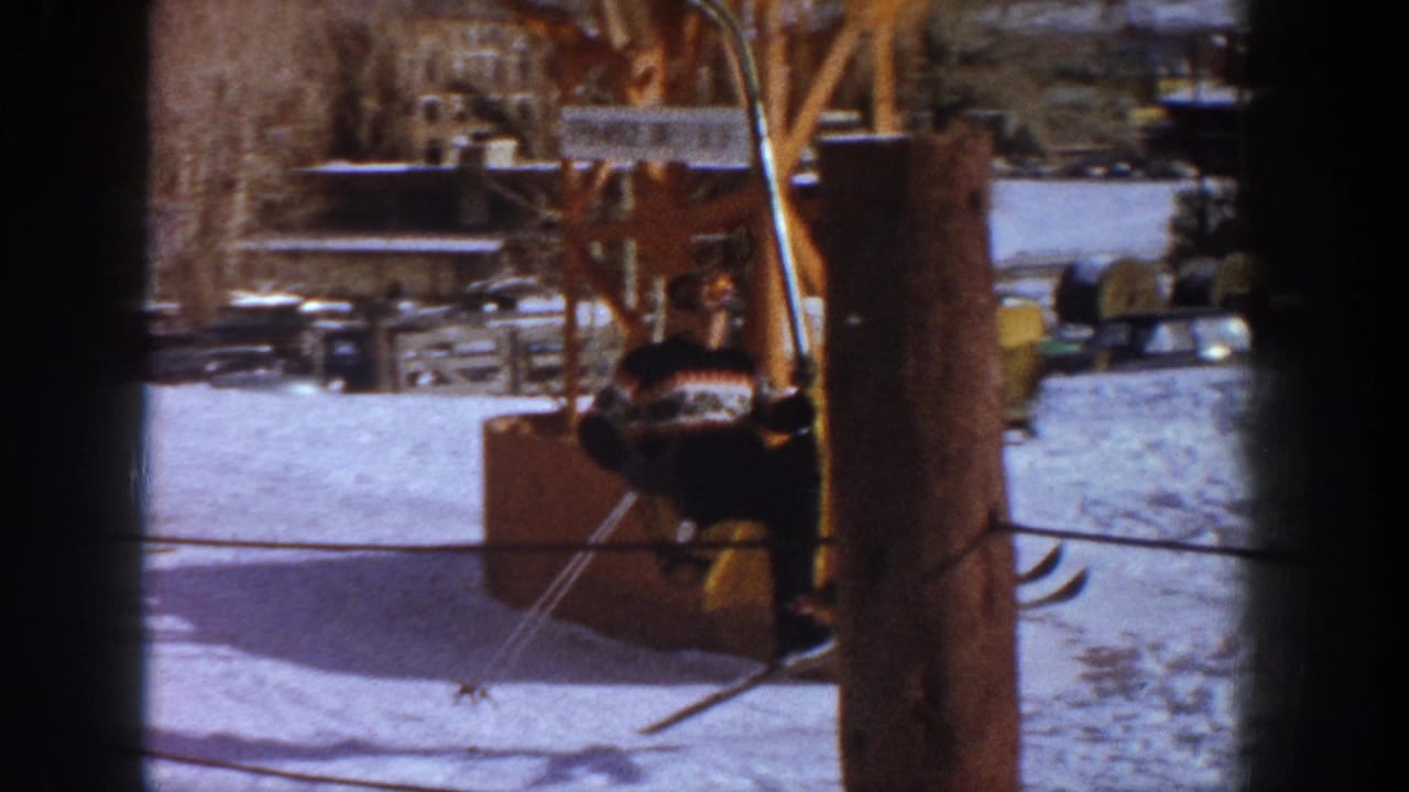 Skier in patterned sweater and yellow pants seated on a wooden ski lift chair, with lift machinery and snowy background.