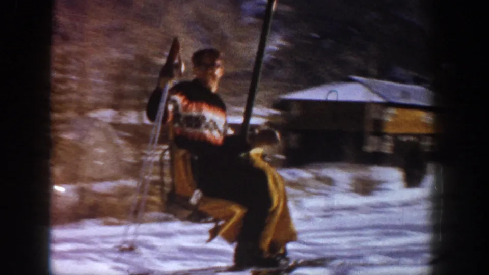 A skier seen from behind in a striped sweater and yellow pants holds ski poles near a wooden lodge at what metadata identifies as Aspen, Colorado, circa 1961.