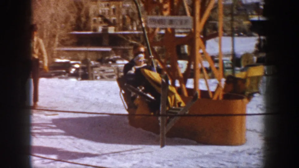 A vintage fixed-grip ski lift loading station with orange-painted wooden machinery at what metadata identifies as Aspen, Colorado, circa 1961; parked period automobiles visible in the background.