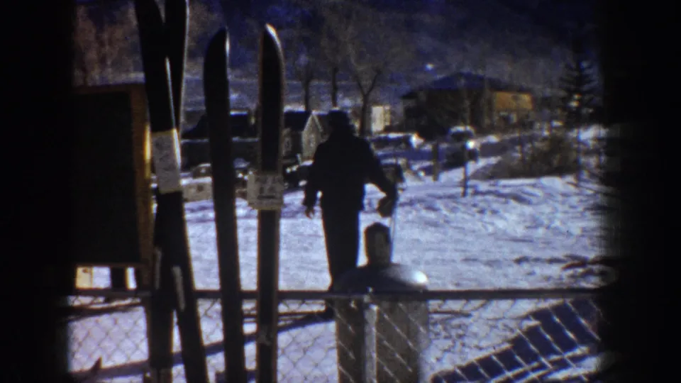 Ski area base, possibly Aspen circa 1961 — skis silhouetted in the foreground, a lone figure crossing toward low structures on snow-covered ground.