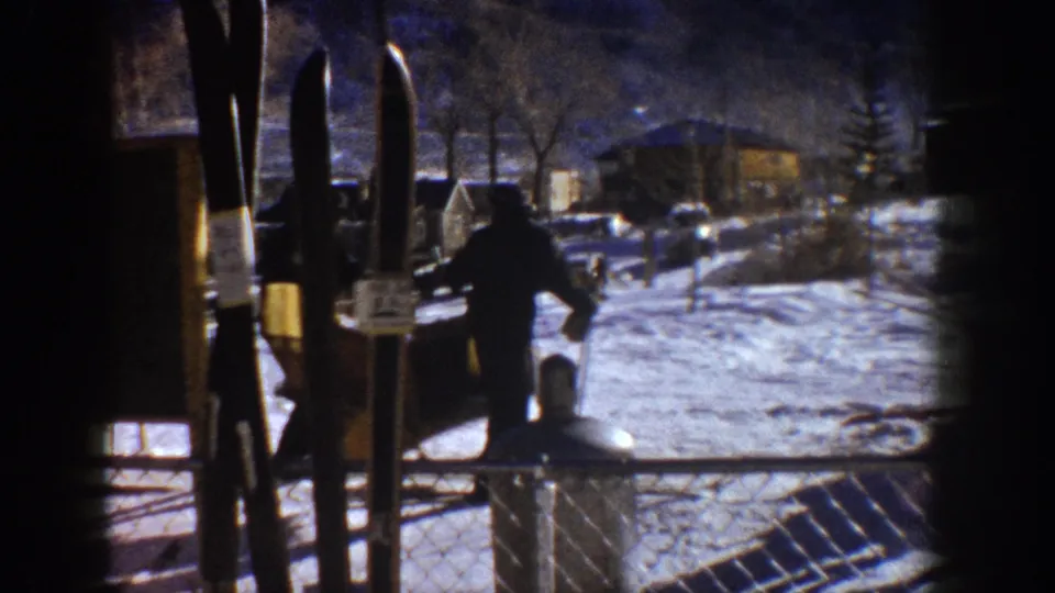 Continuation of the ski base sequence — skis foregrounded, a figure crossing the packed snow at what appears to be an early-1960s Colorado mountain resort.