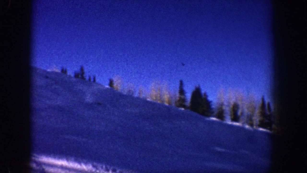 8mm home movie frame from Aspen, Colorado, circa 1961: snow slope in foreground, bare aspen and conifer treeline at ridge, deep blue sky above.