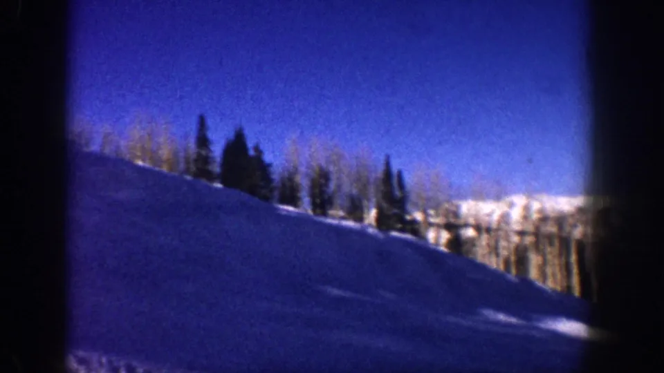 8mm frame from Aspen, Colorado, circa 1961: panning shot shows snow slope, mixed aspen and conifer treeline, saturated blue sky, and edge vignetting.