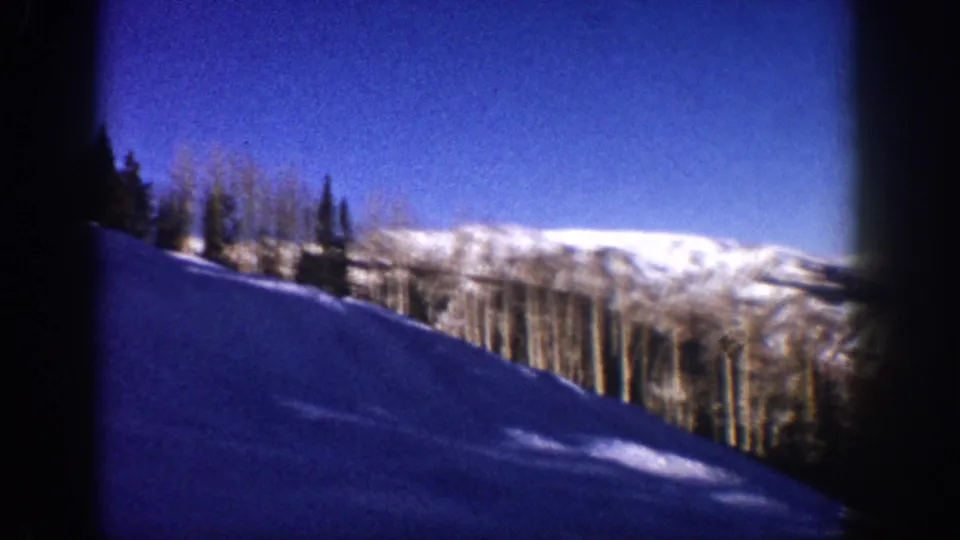 8mm home movie frame from Aspen, Colorado, circa 1961: wide pan reveals snow foreground, aspen and conifer treeline, and bright distant mountain ridge under clear blue sky.
