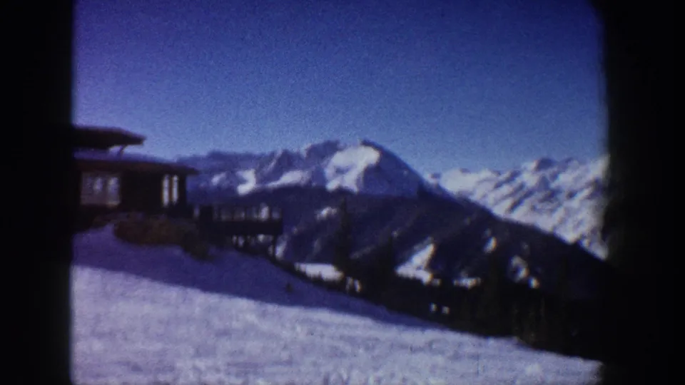 8mm color film frame of a mid-century alpine cabin on a snowy hillside with dramatic snow-capped peaks and vivid blue sky, Aspen, Colorado area, circa 1961.