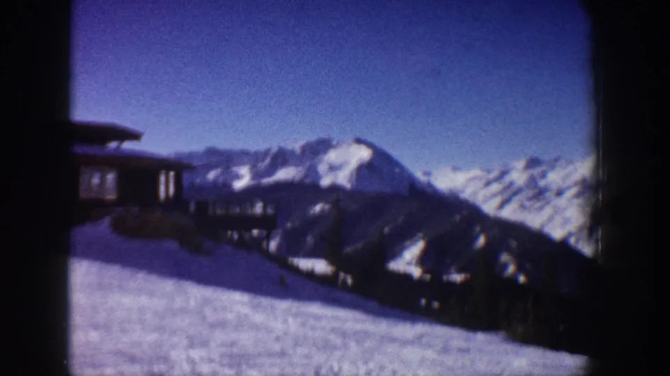 8mm home movie frame of a mid-century alpine cabin on a snowy slope with prominent mountain peaks under a clear blue sky, Aspen, Colorado area, circa 1961.