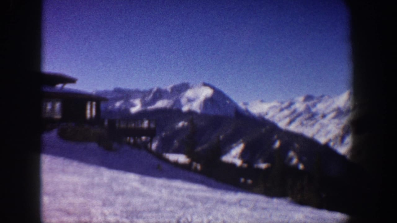 8mm home-movie frame circa 1961 showing snow-covered Aspen-area peaks, blue sky, and a dark wooden lodge structure at upper left.
