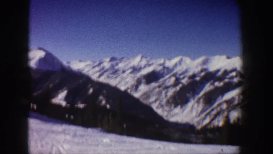 8mm frame circa 1961 showing a broad snow-covered Aspen-area mountain range with conifer trees on mid-slopes and blue sky; lodge no longer in frame.