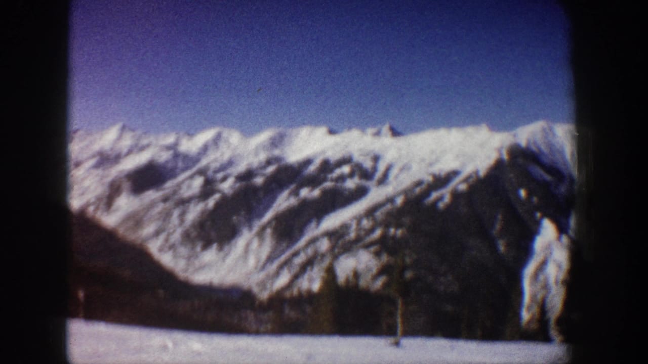 Snow-covered mountain range with tall dark conifer and possible structure at lower right, 8mm film
