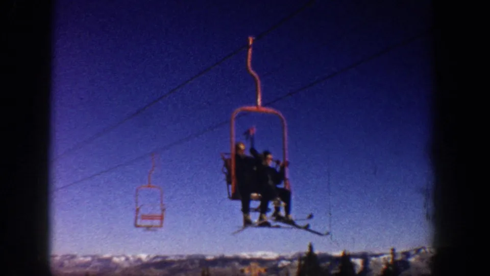 8mm frame circa 1961 of two skiers on a red open-bench chairlift at what metadata identifies as Aspen, Colorado; one rider waves toward the camera.