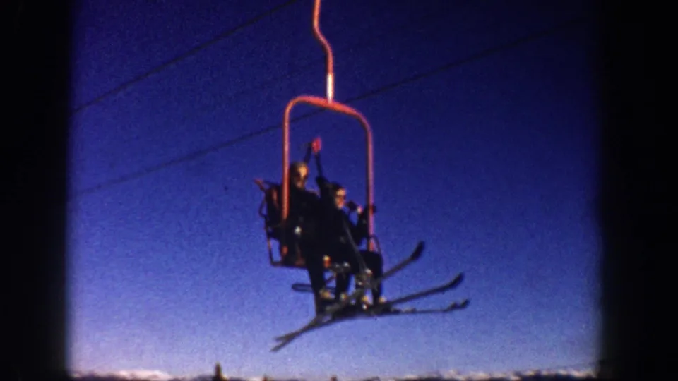 Closer 8mm frame circa 1961 of two skiers on a red open-bench chairlift at what metadata identifies as Aspen, Colorado; faces are partially discernible.