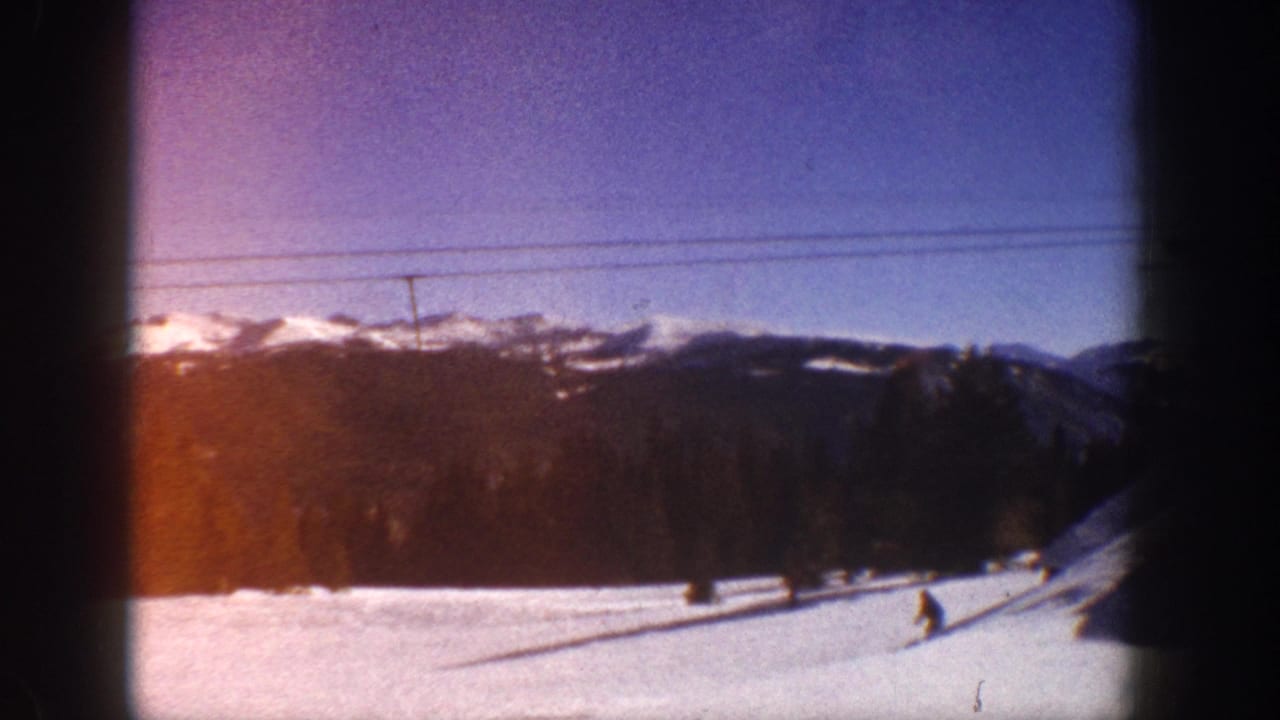Wide shot of a snow-covered ski slope with a dark conifer ridgeline, snow-capped peaks, overhead lift cables, and a distant skier figure.
