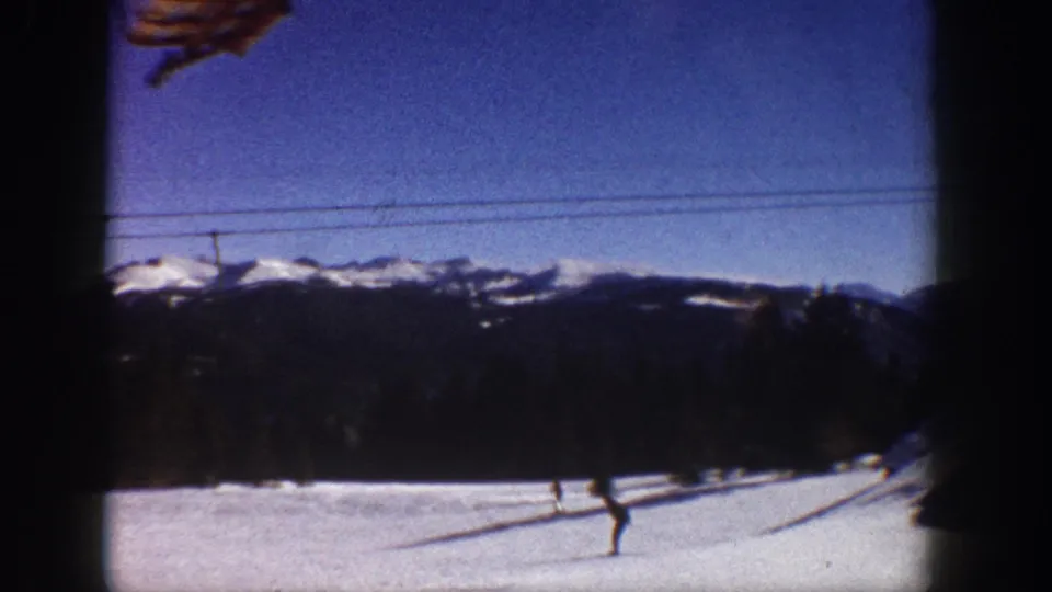 Broad snowfield looking toward a Rocky Mountain ridgeline under a deep blue sky, with lift cables and a lone skier; consistent with Aspen, Colorado, circa 1961 8mm footage.
