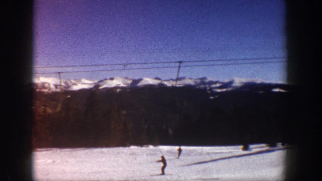 Two skiers on a wide snow slope at what metadata identifies as Aspen, Colorado, circa 1961, with Rocky Mountain peaks and ski lift cables visible.