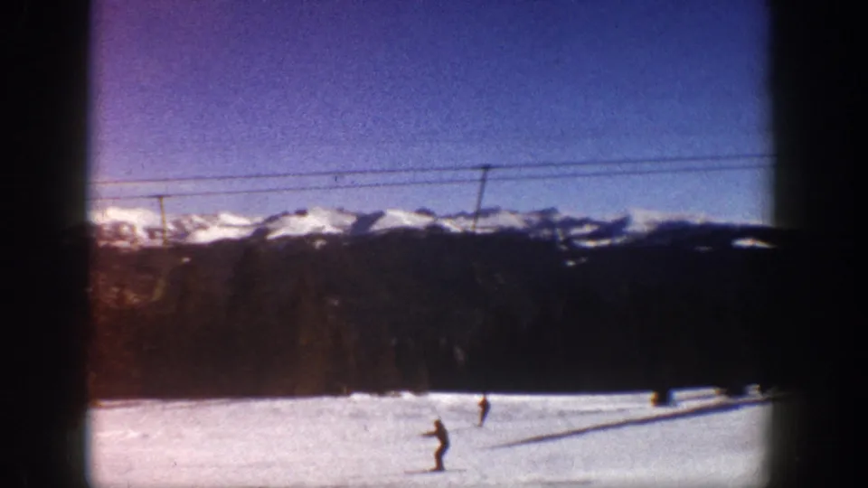 Two skiers on a wide snow slope at what metadata identifies as Aspen, Colorado, circa 1961, with Rocky Mountain peaks and ski lift cables visible.