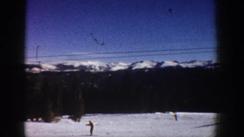 Wide 8mm shot of a ski run with a skier at lower frame, lift cables, and a snow-capped mountain backdrop — Aspen, Colorado circa 1961 per metadata; a film scratch is visible in the upper sky.