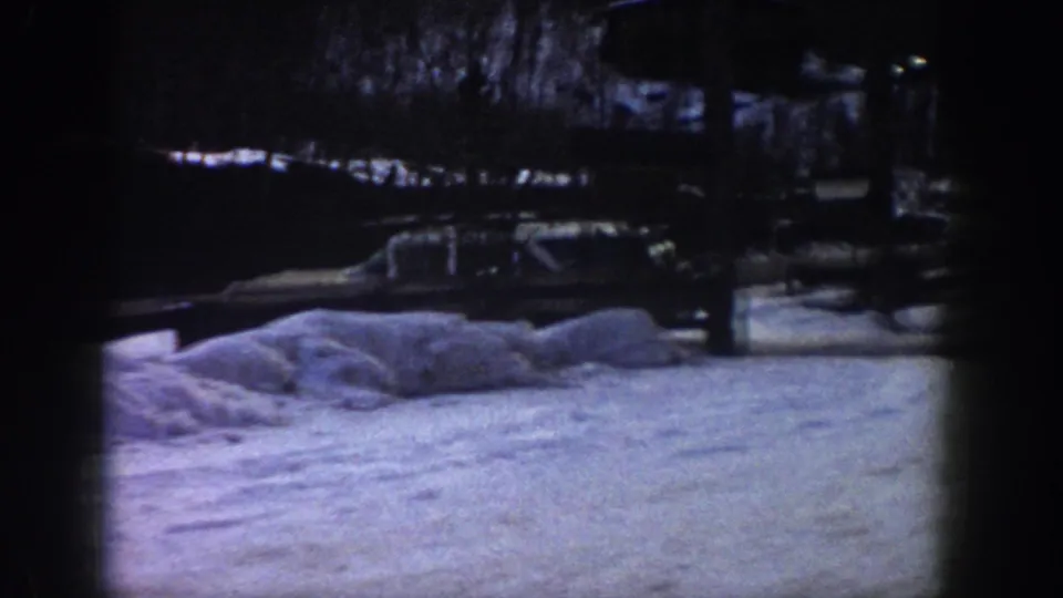 A station wagon on a snowy road through a winter forest with background structures, consistent with an Aspen, Colorado area road scene circa 1961 per clip metadata.
