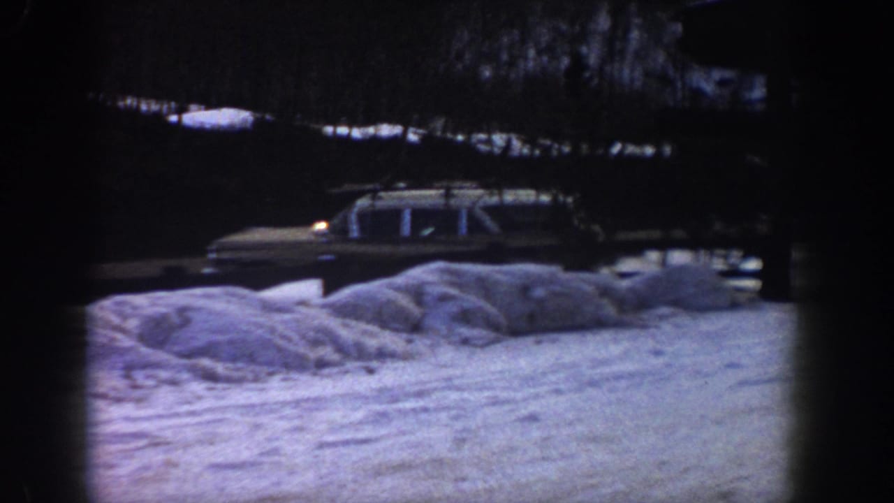 Station wagon with roof rack driving along a snow-covered road, large snow drifts in foreground, blue-purple 8mm color cast.