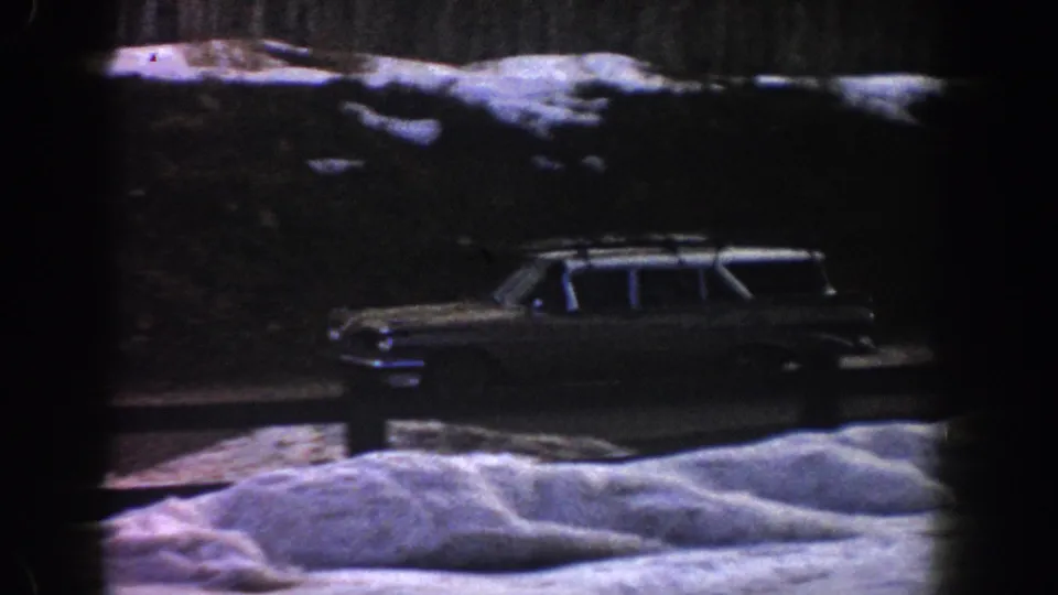 Early-1960s American station wagon with roof rack on a snowy mountain road, likely Aspen, Colorado, 1961, filmed on 8mm with blue-purple color shift.