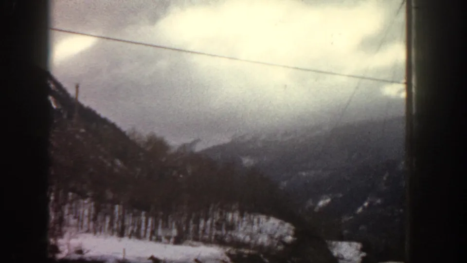 Snow-dusted mountain slope viewed through a vehicle window, utility lines overhead and fence posts at the lower edge; appears to be a Rocky Mountain valley, circa 1961 8mm home movie.