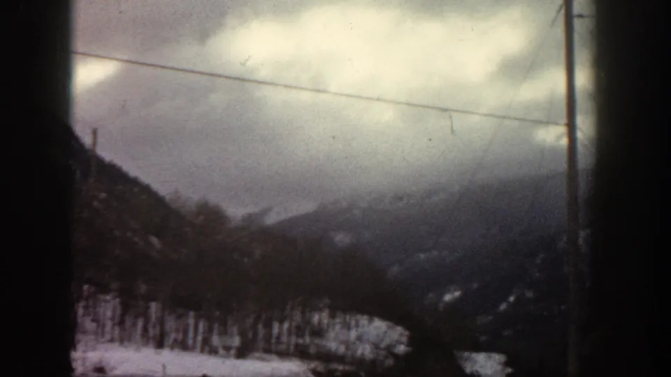 Snow-covered mountain ridge under a heavy overcast sky, utility pole and crossing wires visible, shot through a vehicle window; Rocky Mountain area, circa 1961 8mm film.
