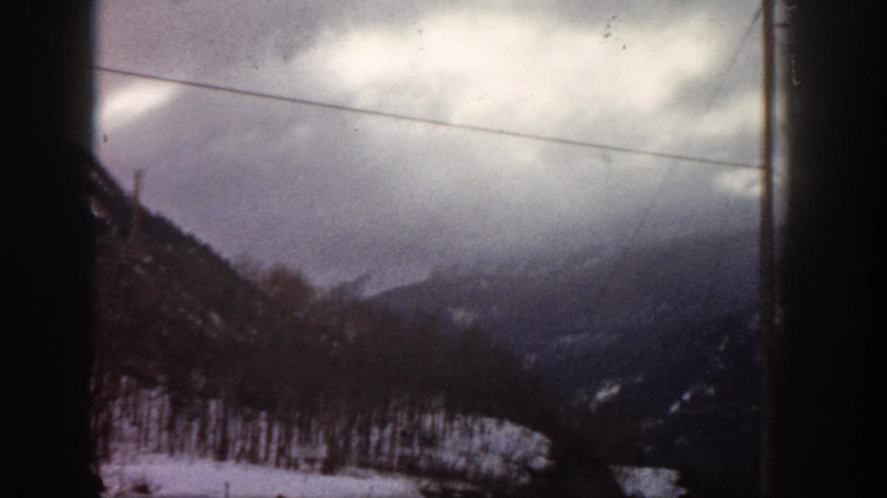 Mountain hillside with snow, power lines, and heavy cloud cover, filmed through a car window on 8mm.