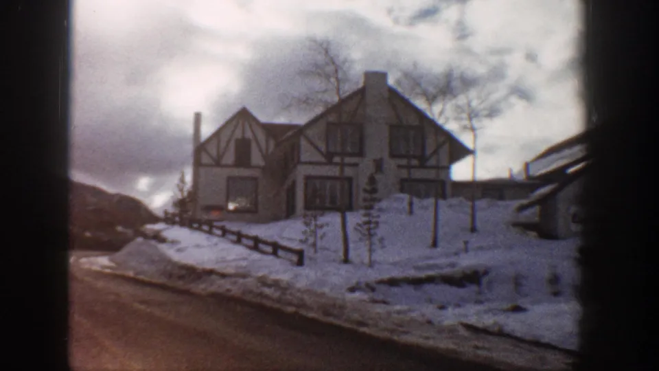 Wide shot from vehicle of Tudor Revival house along a snow-lined paved road, Aspen Colorado, 1961; bare deciduous trees and split-rail fence visible.