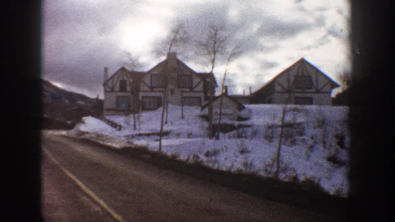8mm home movie frame showing a winding road and a Tudor-style building with snow-covered ground and mountains, likely Aspen, Colorado, circa 1961.